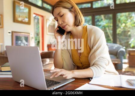 Asian woman talking on smartphone and using laptop, working from home Stock Photo