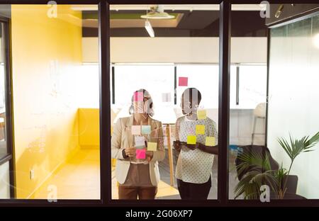 Two diverse businesswomen standing at transparent board, making notes and talking Stock Photo