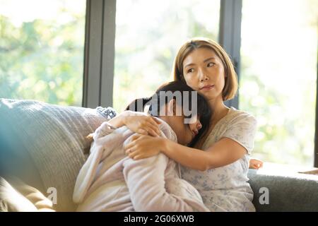 Portrait of serious asian woman with her daughter embracing sitting on sofa Stock Photo