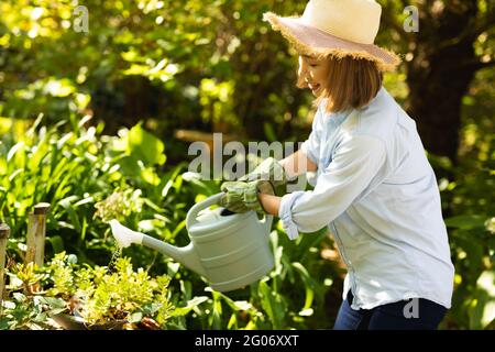 Smiling asian woman wearing straw hat and using watering can in garden Stock Photo