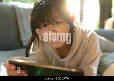 Smiling asian girl in glasses using smartphone and lying on sofa Stock Photo