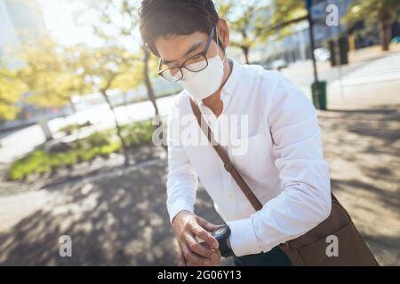 Asian businessman wearing face mask using smartwatch in city street Stock Photo