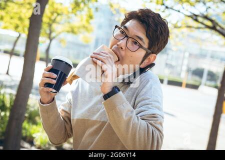 Asian businessman using eating sandwich and holding takeaway coffee sitting in city street Stock Photo