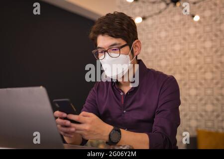 Asian businessman wearing face mask using smartphone and laptop in hotel lobby Stock Photo