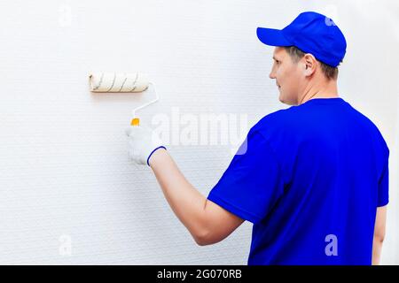 A painter in a cap and gloves paints the wall with a paint roller. Portrait of a young worker in uniform. Professional repair of premises. Mockup Stock Photo