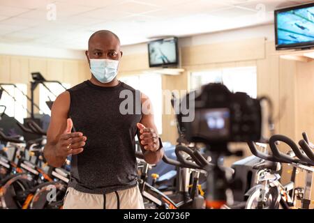 Male sports instructor in gym with a face mask recording video content for online classes, streaming... Behind the scenes. Selective focus. Stock Photo