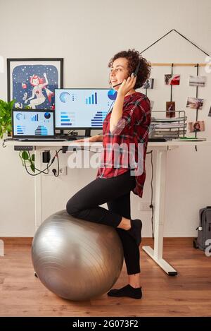 Woman teleworking at an adjustable standing desk with one knee resting on a fitball Stock Photo