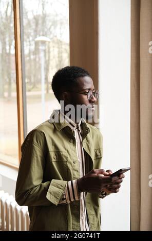 African businessman in eyeglasses looking through the window and using mobile phone while standing at office Stock Photo
