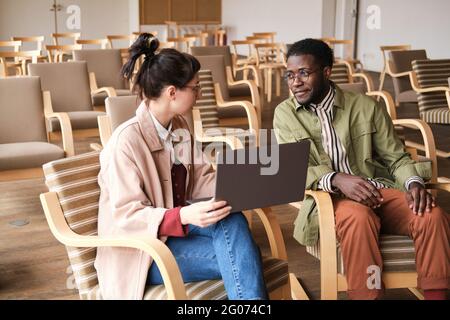 Young businesswoman using laptop and talking to her colleague while they sitting in empty conference hall Stock Photo