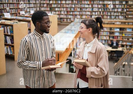 Young happy couple with books talking to each other while standing in the library Stock Photo