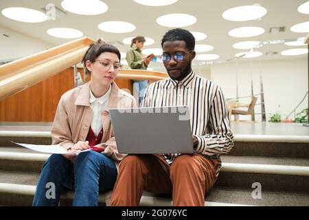 African man using laptop and studying together with woman while they sitting on stairs in the library Stock Photo
