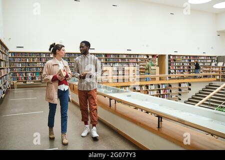 Young multiethnic couple with books talking to each other while walking along the corridor in the library Stock Photo