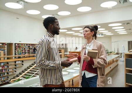 Two students holding books and discussing something while studying in the library Stock Photo