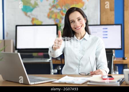Young smiling businesswoman showing thumbs up gesture while sitting at her working table in office Stock Photo