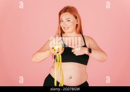 Portrait of beautiful oversized woman wearing sports outfit holding apple with measuring tape, pointing with finger, smiling while looking at camera. Stock Photo