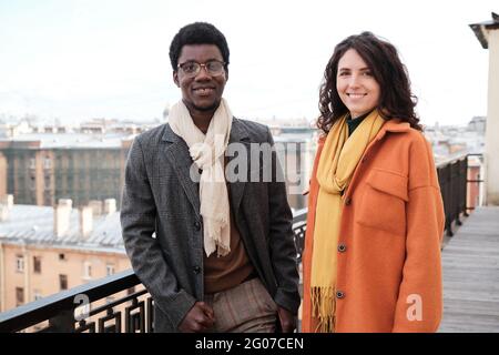 Portrait of multiethnic couple in warm coats smiling at camera while standing on balcony Stock Photo