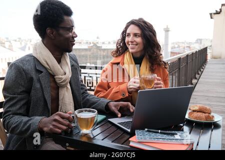 Young couple sitting at the table with laptop and talking during breakfast in outdoor cafe Stock Photo