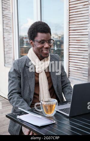 African man sitting at the table in front of laptop and talking online while sitting in outdoor cafe Stock Photo