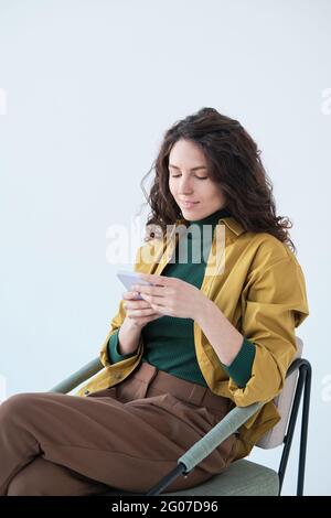 Young beautiful woman sitting on chair and reading online on the phone isolated on white background Stock Photo