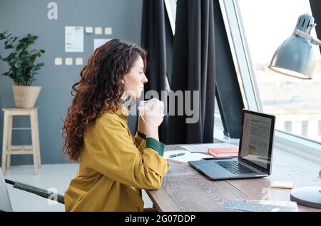 Young woman drinking coffee while sitting at the table in front of laptop and working at home Stock Photo
