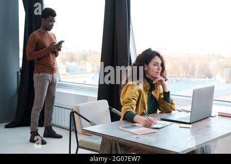 Young businesswoman working on laptop at the table with man standing behind her and using mobile phone Stock Photo