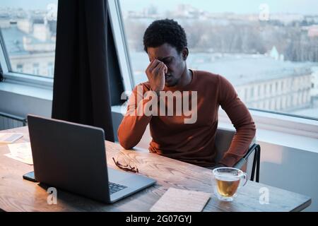 African businessman sitting at the table in front of laptop and looking very tired Stock Photo