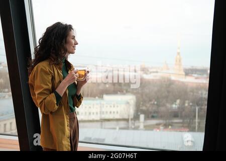 Young businesswoman holding cup of tea and looking through the window at city while standing at office Stock Photo
