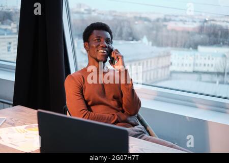 African smiling businessman talking on mobile phone while sitting at the table in front of laptop at office Stock Photo