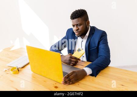 Pensive young Afro American businessman sit at desk in office work on laptop solving business problem. Serious male employee holding smart phone and Stock Photo
