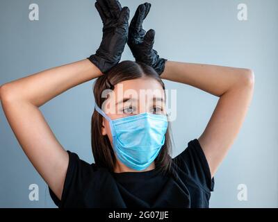Female teenage girl in a blue protective medical face mask shows corona by holding her hands in black latex gloves above her head. Coronavirus concept