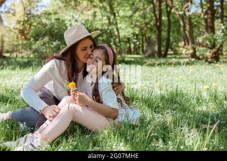 Mother kissing her happy daughter sitting on grass in summer park. Family relaxing outdoors picking wild flowers Stock Photo