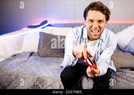 Joyful young Caucasian male playing game at home on console station. Smiling millennial man gamer having fun enjoy world of videogames, cyber sport Stock Photo