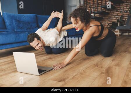 Fit young man doing sport exercise at home with laptop lying on floor with girlfriend supporting helping boyfriend, sporty female training husband at Stock Photo