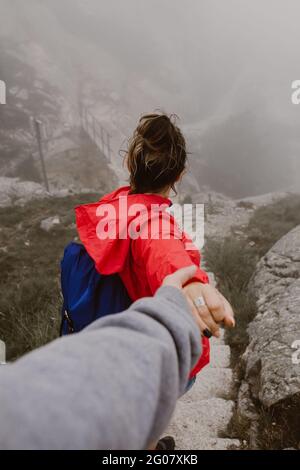 Back view of young tourist with backpack holding hand of human and walking on hill near footbridge Stock Photo