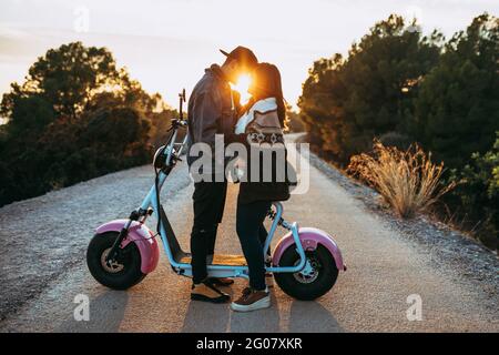 Side view of young hipsters embracing near vintage scooter on countryside route at sunset Stock Photo