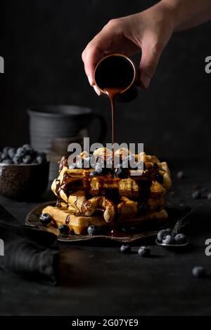 Crop anonymous person adding chocolate syrup from small cup on top of delicious waffles pyramid with blueberries on kitchen Stock Photo