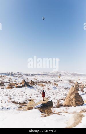 Full body unrecognizable female tourist standing on stone and admiring hot air balloons flying in cloudless blue sky over snowy mountainous terrain on Stock Photo