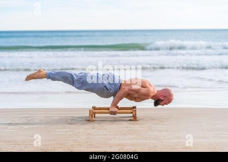 Side view of bald strong athlete doing parallel handstand with bars working out on sandy coast Stock Photo
