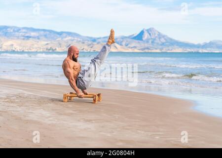 Side view of focused bald strong athlete working out lifting up his own wait from bars on sandy coast Stock Photo