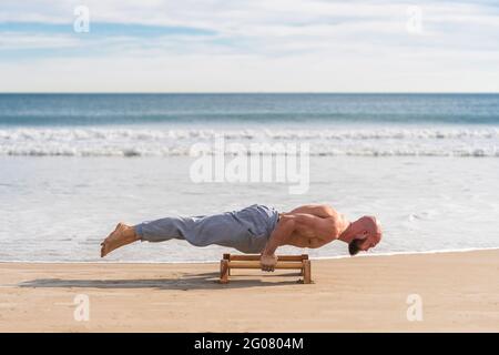 Side view of bald strong athlete doing parallel handstand with bars working out on sandy coast Stock Photo