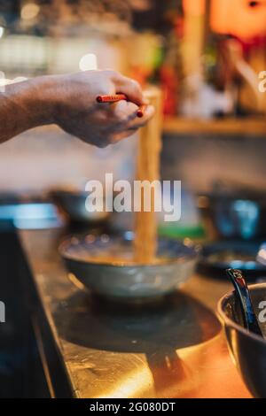Crop hand of faceless chef with wooden chopsticks holding noodle above bowl on blurred background in ramen bar Stock Photo