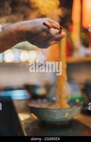 Crop hand of faceless chef with wooden chopsticks holding noodle above bowl on blurred background in ramen bar Stock Photo