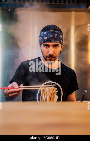 Young bearded male chef in bandana looking at camera while holding noodle with chopsticks in ramen bar Stock Photo