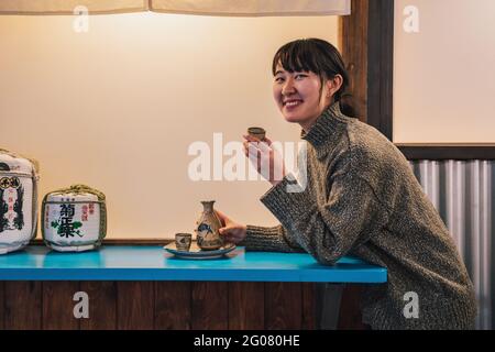 Side view of overjoyed Asian Woman in sweater sitting at blue table with traditional drink in cafe Stock Photo