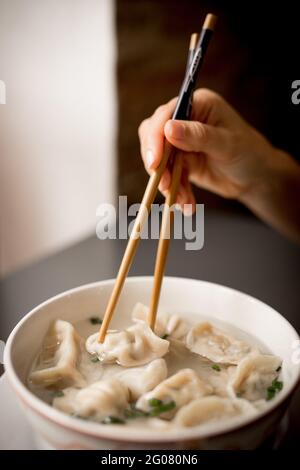 Hand of Woman with traditional asian chopsticks eating dumpling soup in ceramic bowl Stock Photo