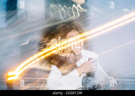 Unrecognizable cheerful black guy in stylish outfit giving piggyback ride to happy girlfriend taking selfie on mobile phone near freeze lights on stre Stock Photo