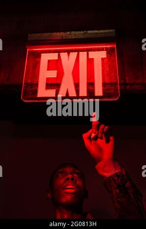 From below crop African American young male looking up pointing at illuminated tablet Exit above head in red dark light Stock Photo