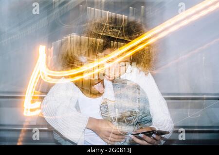 Unrecognizable cheerful black guy in stylish outfit giving piggyback ride to happy girlfriend browsing on mobile phone near freeze lights on street Stock Photo