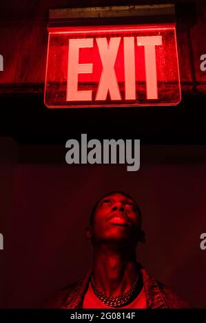 From below crop African American young male looking up on illuminated tablet Exit above head in red dark light Stock Photo