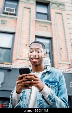 From below black positive young man in jeans outfit messaging on mobile phone while walking in city Stock Photo